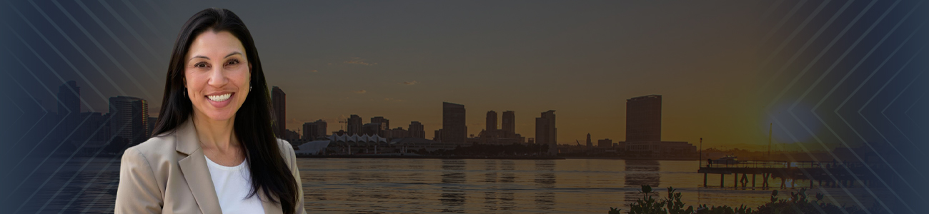 A woman in a beige coat and white shirt smiles while standing by a lake with tall buildings and a dock in the background. The sky is clear and sunny with a beautiful sunset.