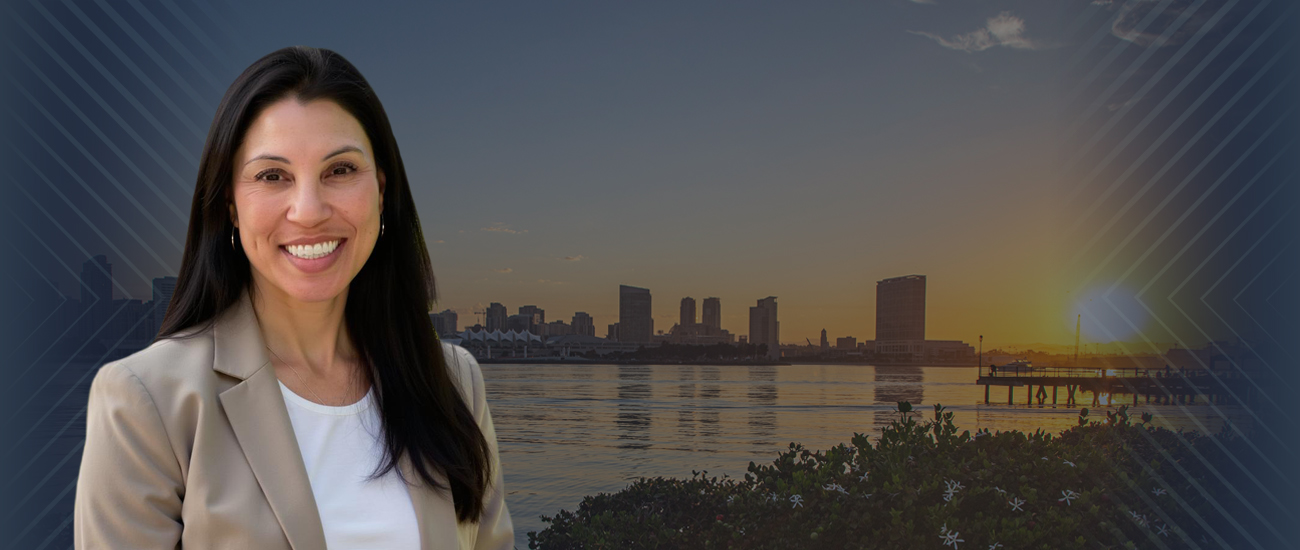 A woman in a beige coat and white shirt smiles while standing by a lake with tall buildings and a dock in the background. The sky is clear and sunny with a beautiful sunset.