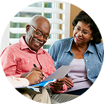 An older African-American couple is sitting on a couch, smiling as they look at a piece of paper.